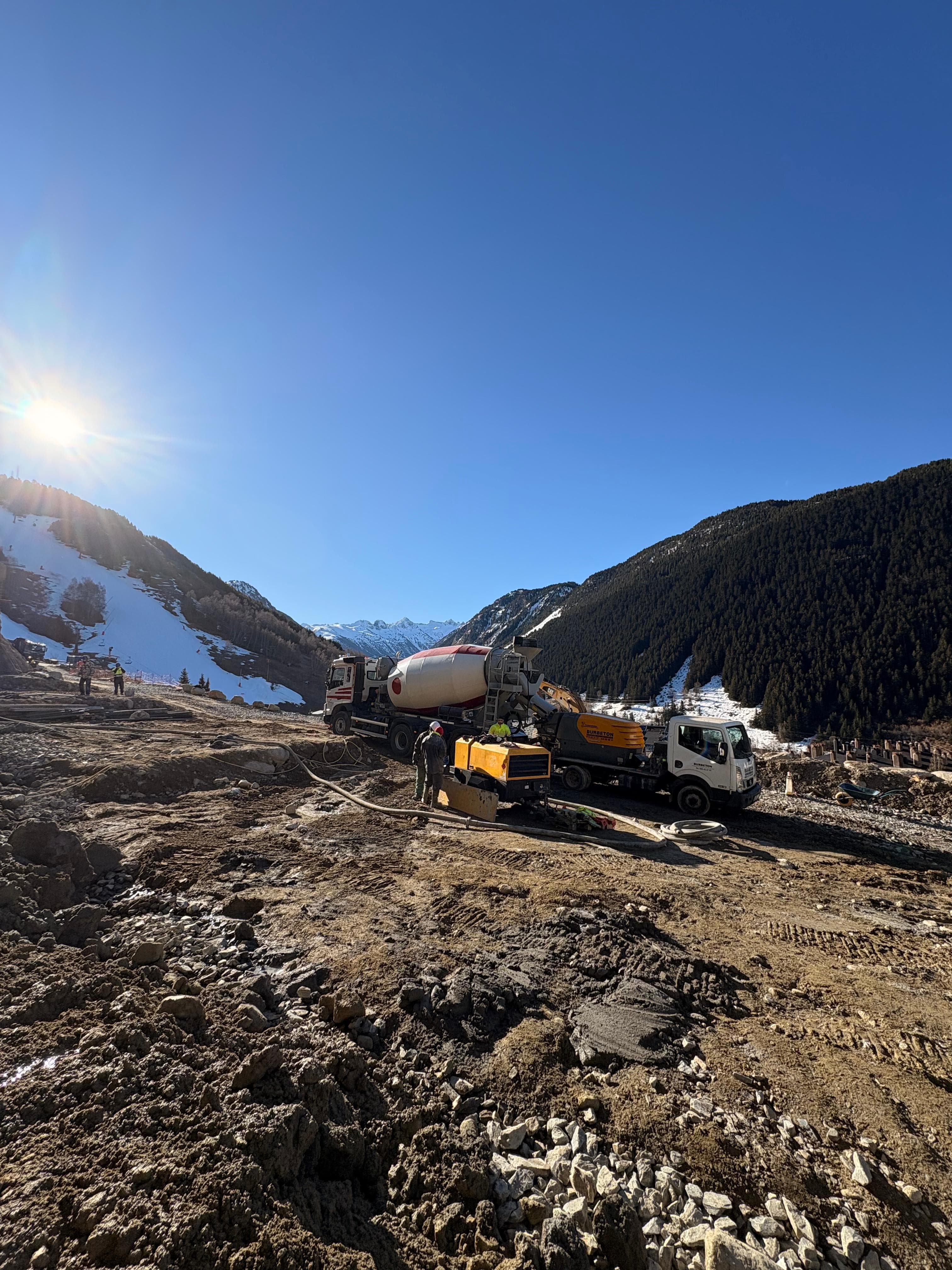 Camión hormigonera y operarios trabajando en una construcción de montaña nevada bajo el sol.