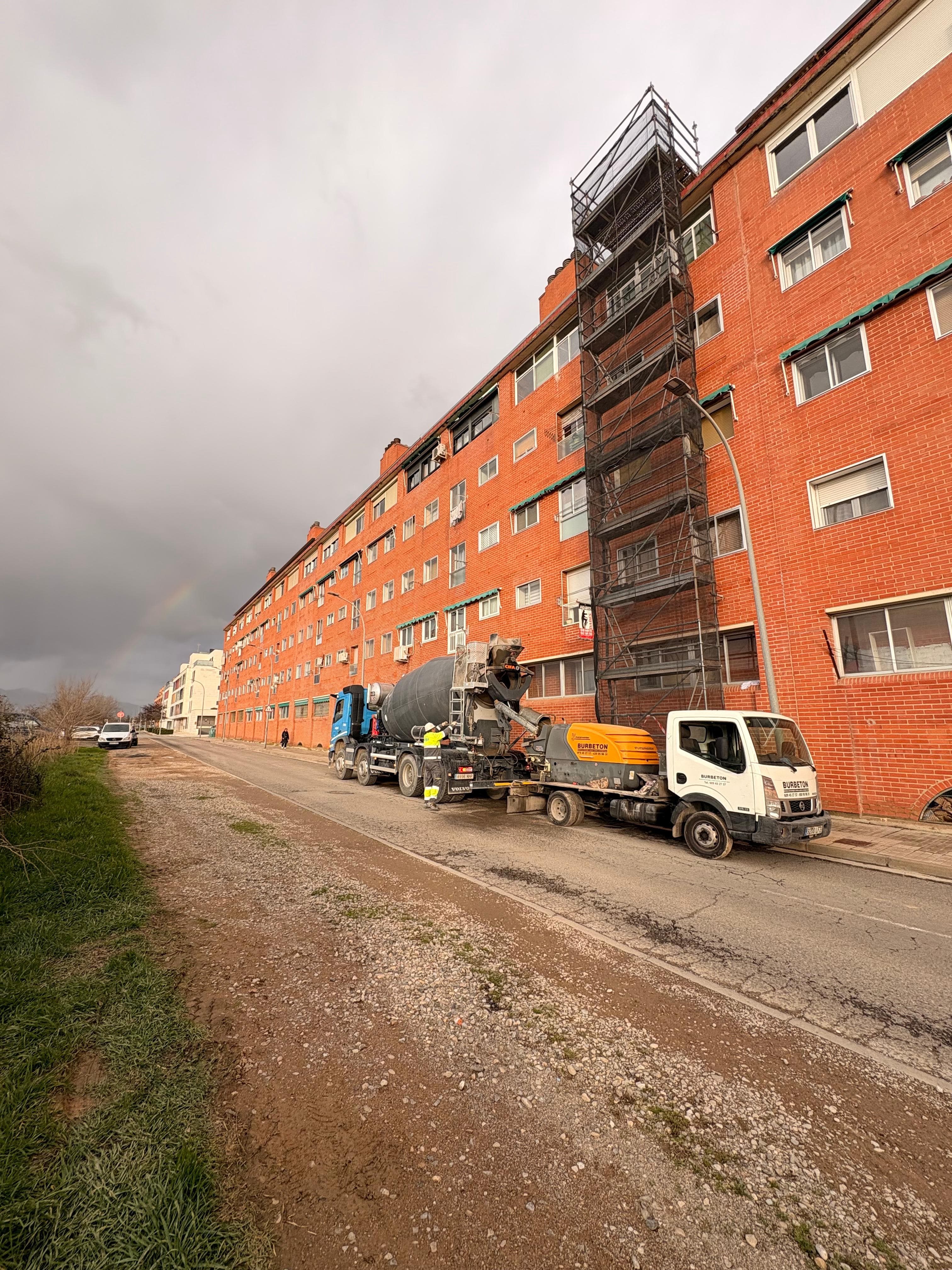 Camión hormigonera y bomba frente a edificio de ladrillo rojo con andamios y arcoíris.