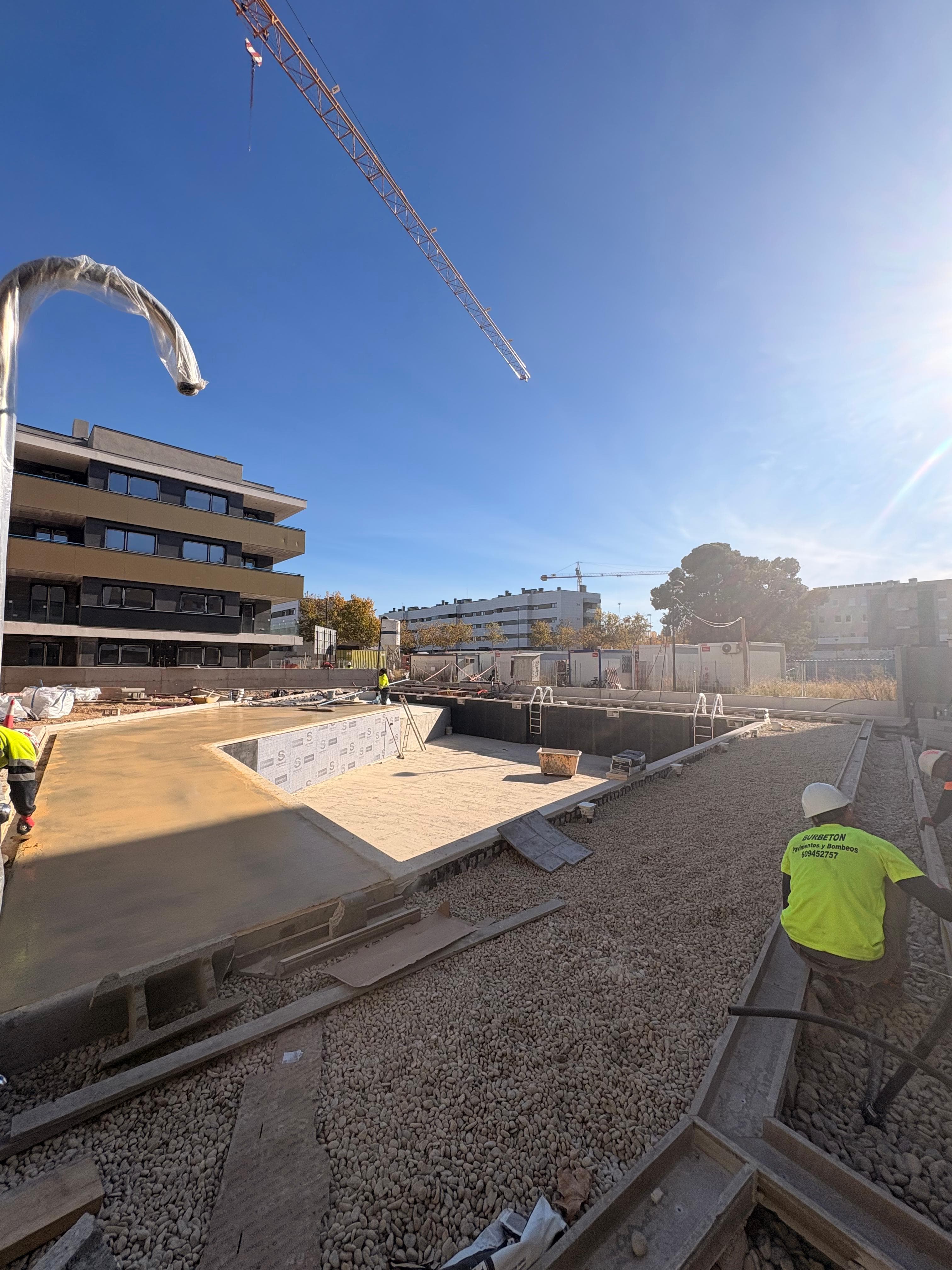 Construcción de una piscina con obreros, grava y una grúa frente a edificios de apartamentos.