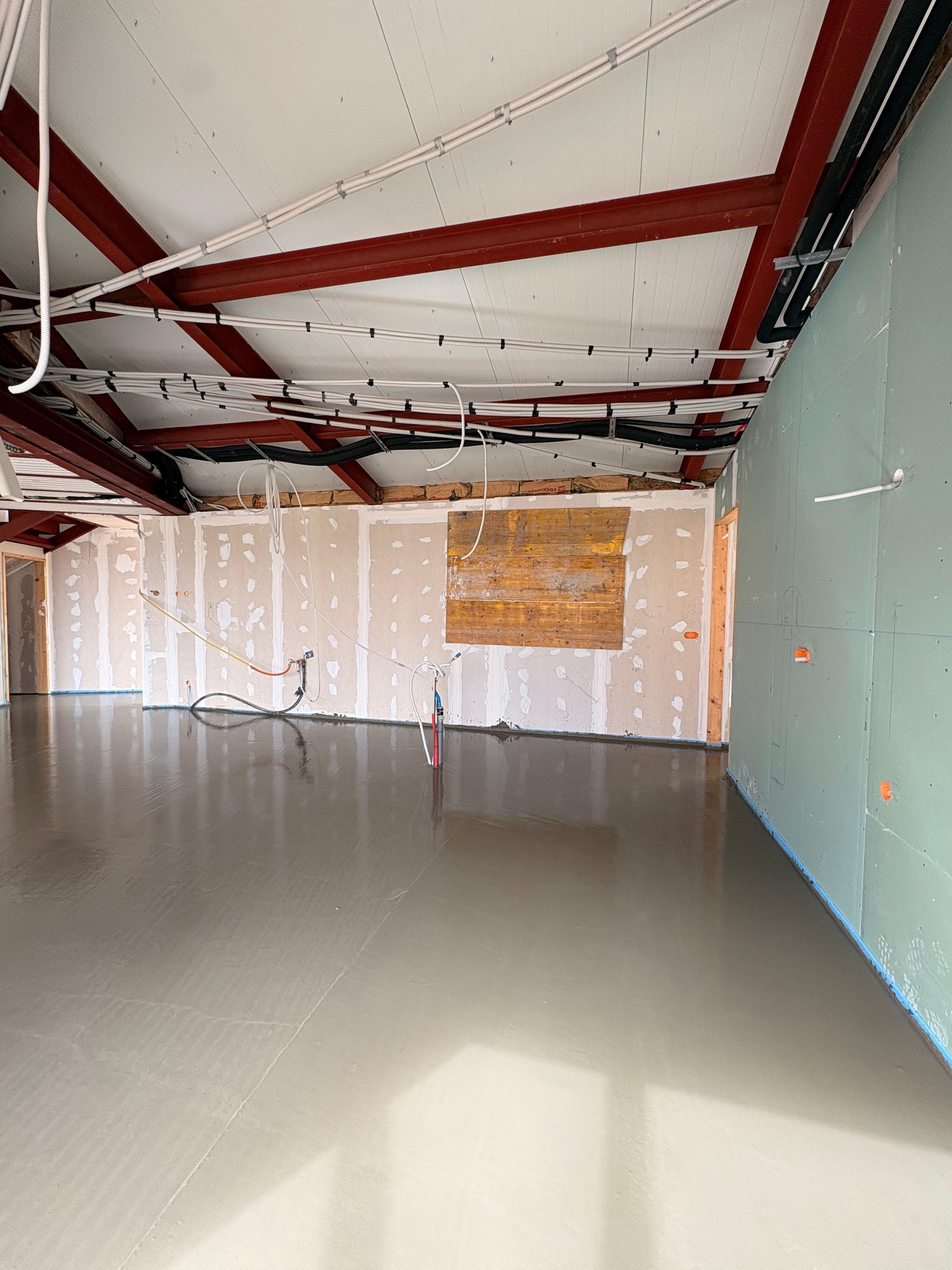Interior en construcción con suelo gris liso, paredes de pladur y vigas de acero rojas.