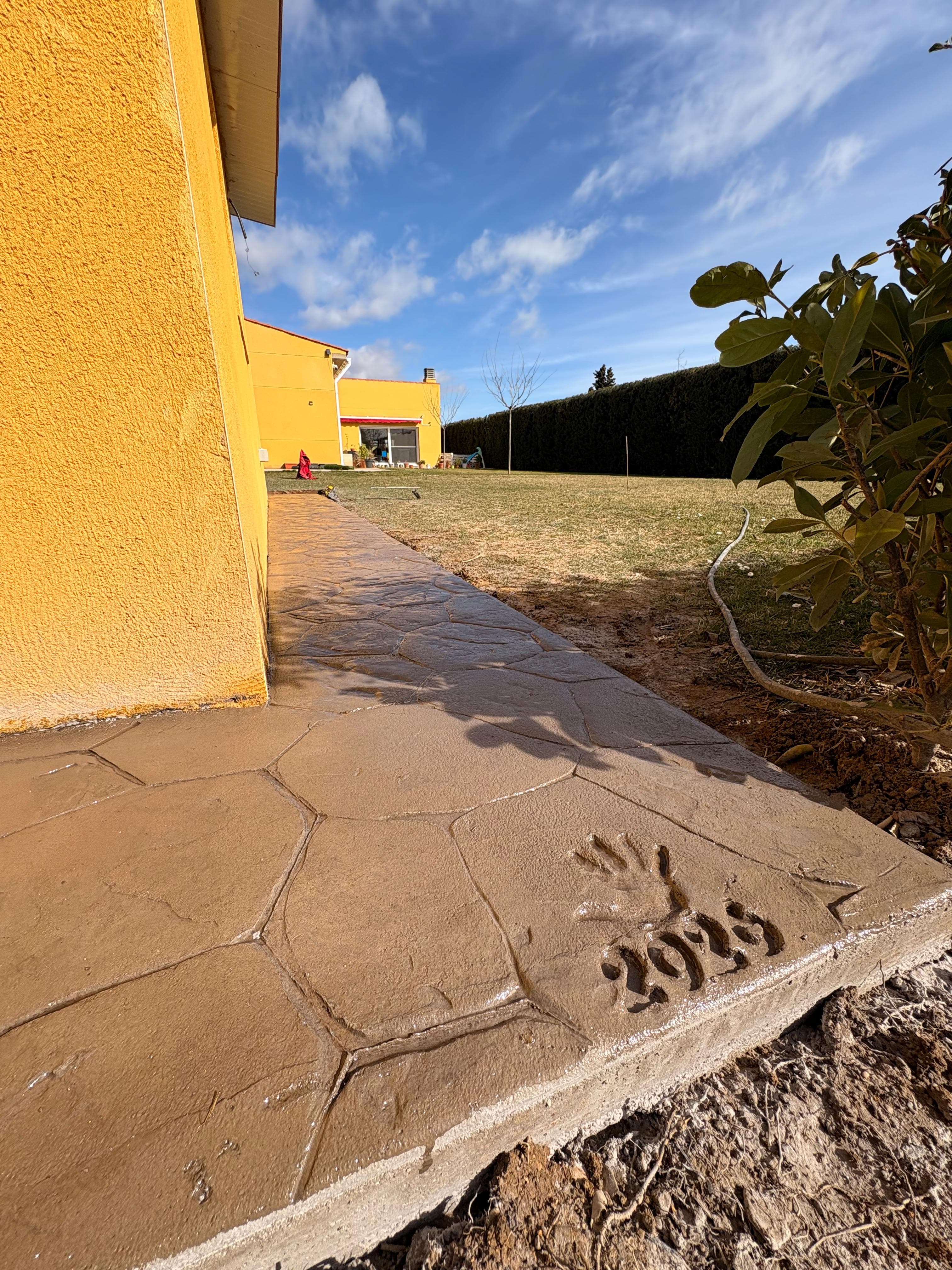 Sendero de hormigón con huella de mano y año 2023 junto a pared amarilla.