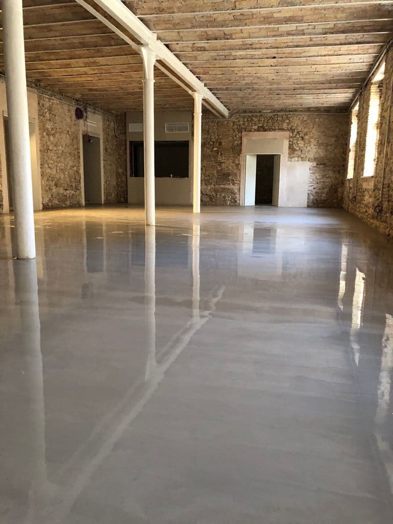 Salón industrial vacío con suelo gris reflectante, paredes de piedra rústica y columnas blancas.
