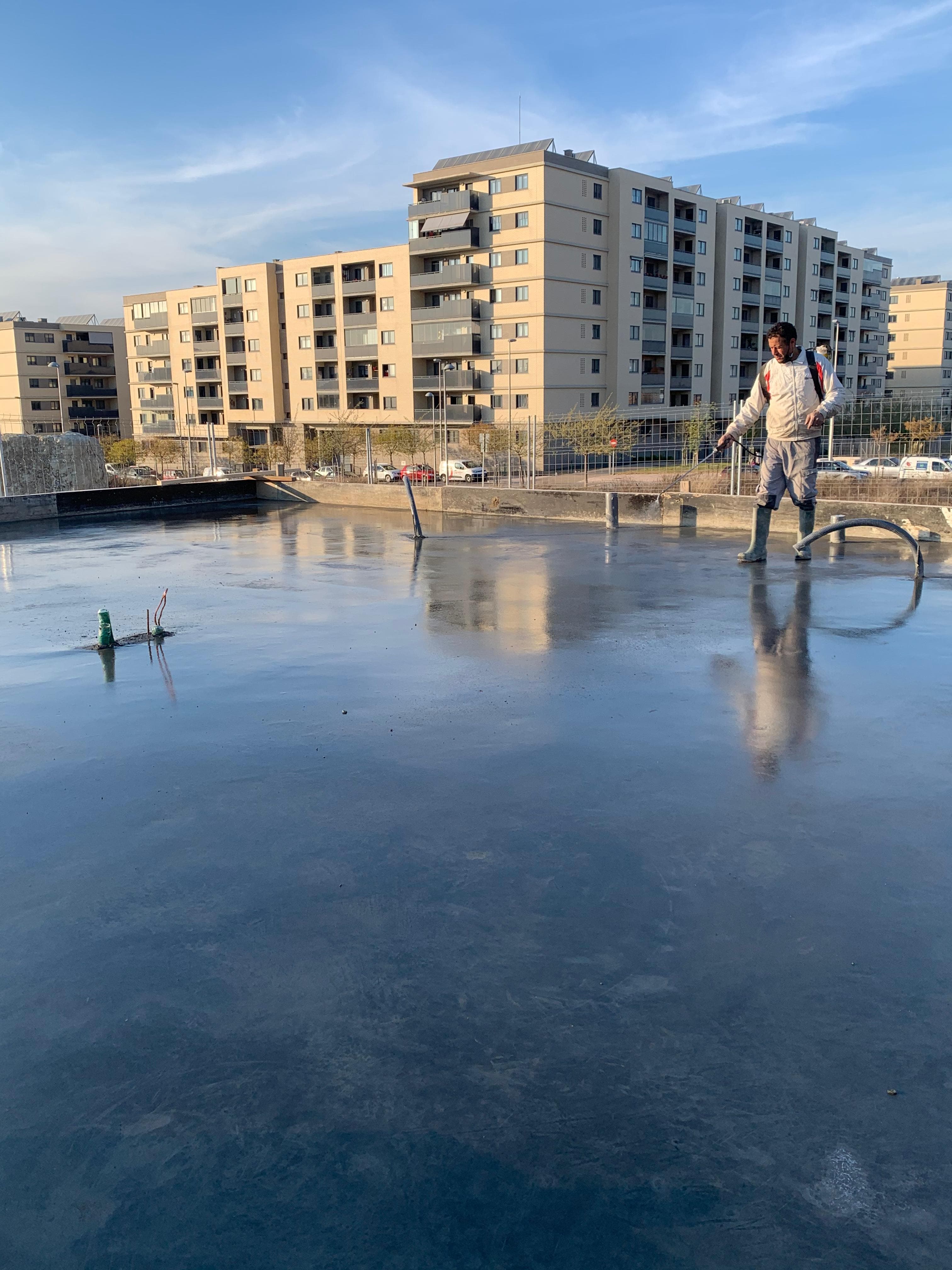 Obrero aplicando impermeabilizante en una azotea húmeda y reflectante con edificios residenciales al fondo.