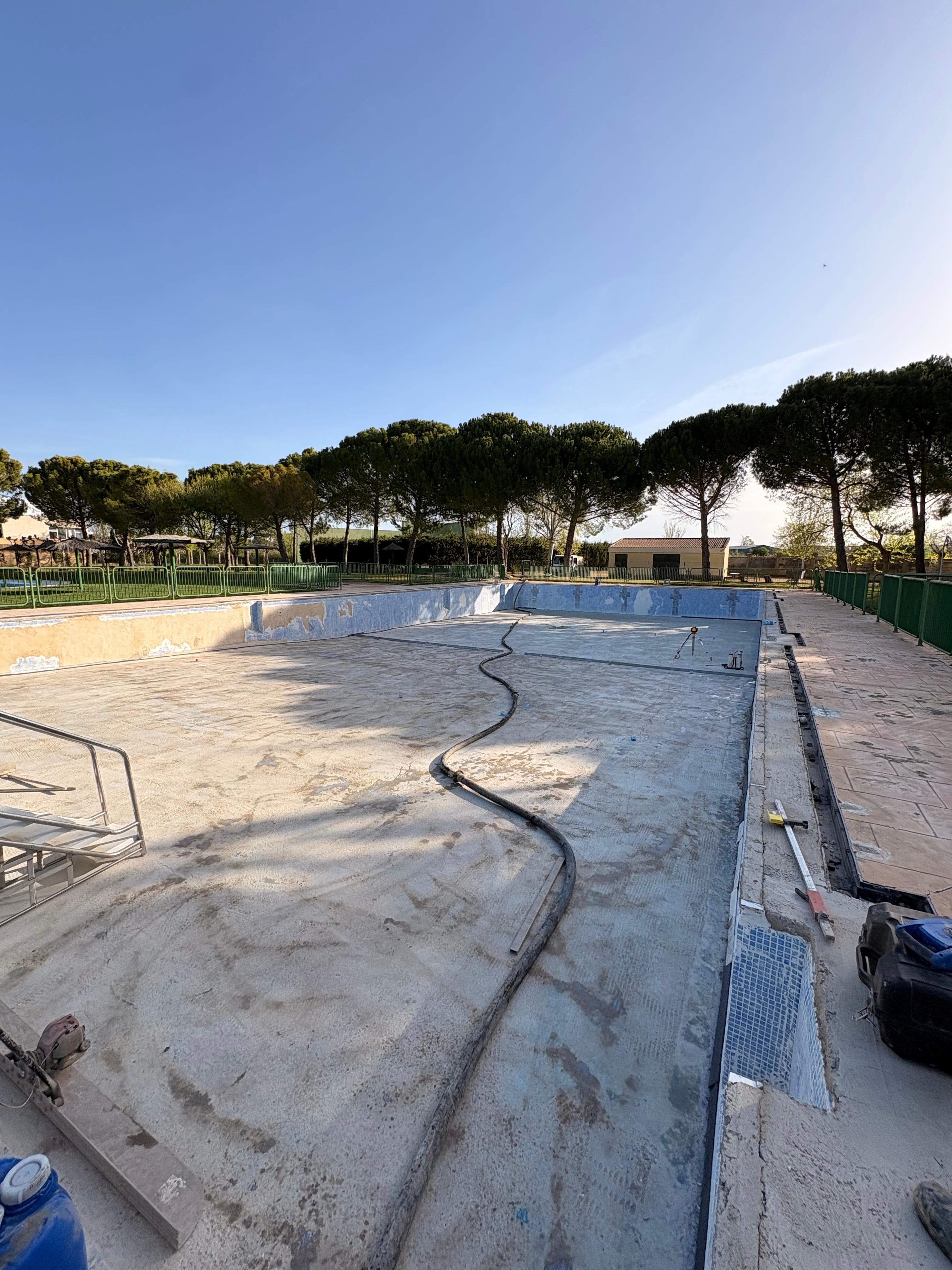Piscina vacía en renovación con manguera sobre el suelo de hormigón y árboles al fondo.